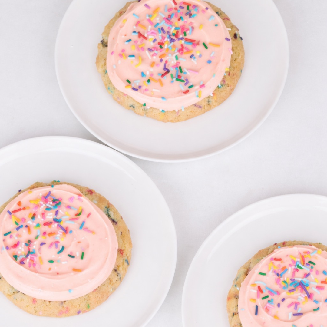 Three cookies with peach frosting and rainbow sprinkles on white plates.