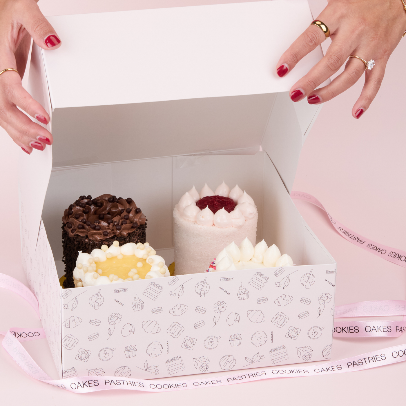 Box of assorted mini 4" cakes with a person's hands opening it, on a pink surface.