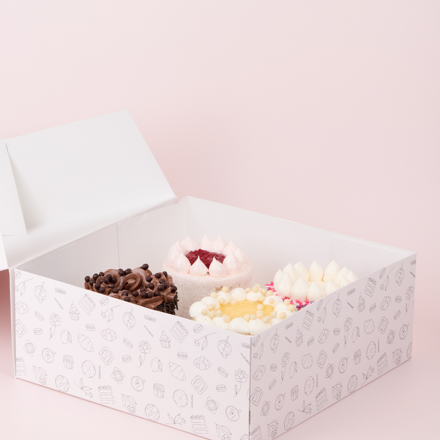 Open box containing three decorated mini cakes on a light pink background